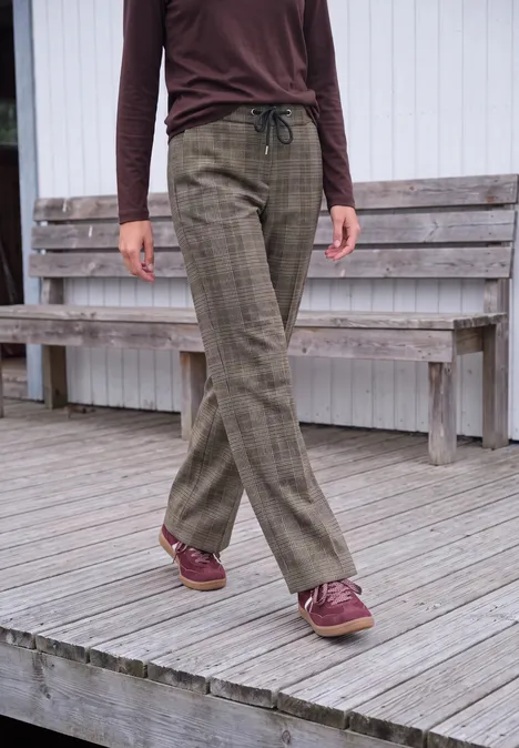 Frau trägt braunes Oberteil, karierte, olivgrüne Hose und weinrote Schuhe auf Holzterrasse.