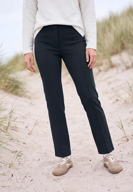 Untere Körperhälfte in schwarzer Hose und beigen Schuhen steht auf sandigem Dünenboden.