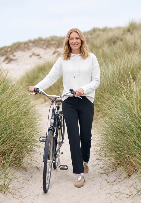 Frau mit Fahrrad läuft durch Dünen am Strand, trägt weißen Pullover und schwarze Hose, lächelnd.