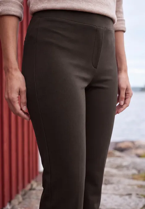 Bootcut-broek met High Waist in Slim Fit truffle brown Vrouw bij een rood houten huisje aan het water, met een donkergrijze, nauwsluitende broek op een stenen pad.