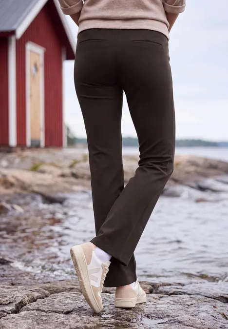 Bootcut-broek met High Waist in Slim Fit truffle brown Persoon in zwarte broek en beige sneakers loopt over rotsige kust bij een rood huis op de achtergrond.