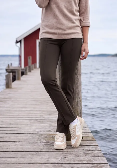 Bootcut-broek met High Waist in Slim Fit truffle brown Persoon op houten steiger aan zee, beige trui, bruine broek en witte sneakers, rood boothuis op achtergrond.
