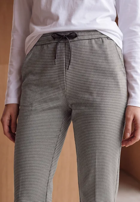 Frau trägt graue Hahnentrittmuster-Hose und weißes Langarmshirt vor holzfarbenem Hintergrund.