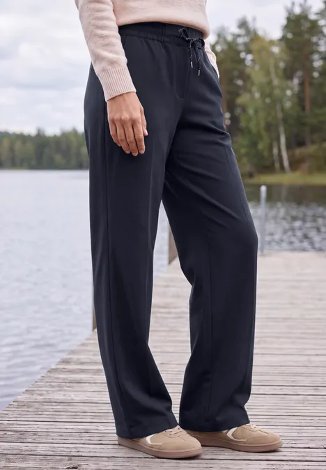 Broek met High Waist en Wide Leg pijpen in een Loose Fit pasvorm urban dark blue Vrouw in ruime zwarte broek en pantoffels staat op een steiger bij een bosmeer.