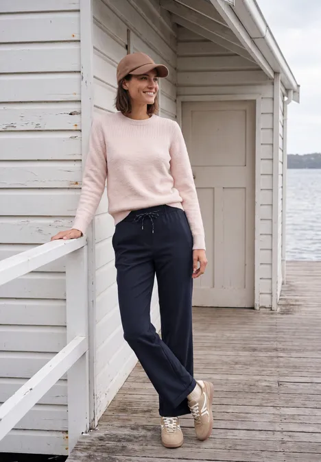 Broek met High Waist en Wide Leg pijpen in een Loose Fit pasvorm urban dark blue Vrouw in roze trui, donkerblauwe broek en pet leunt tegen houten reling voor wit boothuis aan het water.