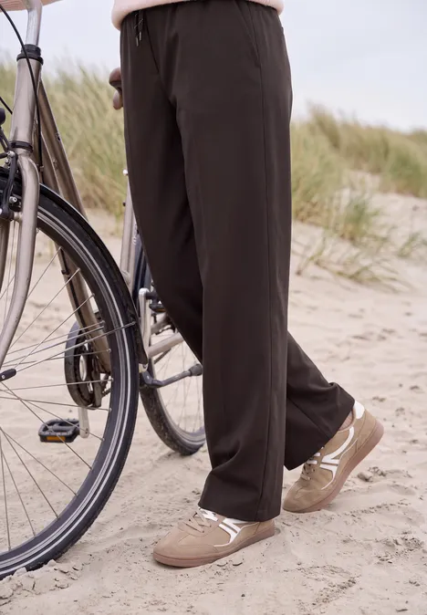 Broek met High Waist en Wide Leg pijpen in een Loose Fit pasvorm truffle brown Persoon in zwarte wijde broek en beige sneakers op een zandstrand naast een fiets.