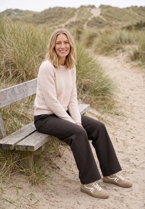 Broek met High Waist en Wide Leg pijpen in een Loose Fit pasvorm truffle brown Een vrouw zit ontspannen op een bankje tussen het helmgras in de duinen en lacht.