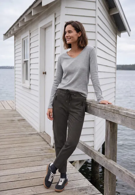 Frau im grauen Shirt und dunkler Hose lehnt lächelnd an einem Steg mit weißer Hütte und Wasser im Hintergrund.