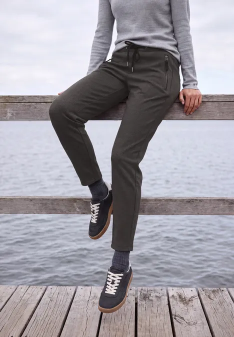 Person lehnt an einem Holzgeländer am Wasser, trägt graue Stoffhose und dunkelblaue Sneaker.