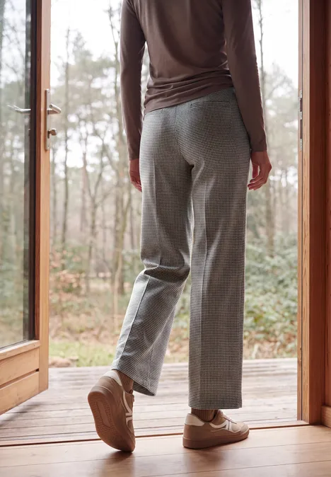 Wide Leg Hose mit Houndstooth-Muster cashmere beige Person steht auf einer Holzterrasse vor einer Glastür, trägt graue Hose und beigen Sneaker, im Hintergrund Bäume.