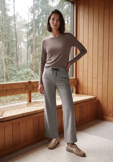 Wide Leg Hose mit Houndstooth-Muster cashmere beige Eine Frau in bequemer Hauskleidung steht in einem Holzraum am Fenster mit Waldsicht.