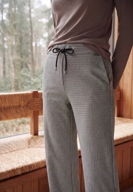 Wide Leg Hose mit Houndstooth-Muster cashmere beige Person sitzt auf Holzbank, trägt graue, karierte Jogginghose mit Tunnelzug und braunes Langarmshirt.