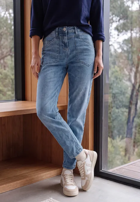 Jean taille mi-haute à Barrel Leg, coupe décontractée mid blue wash Jambes d'une personne en jean bleu et baskets blanches, posée devant une fenêtre cosy avec vue sur la forêt.