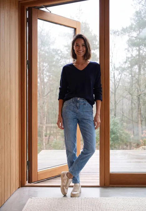 Jean taille mi-haute à Barrel Leg, coupe décontractée mid blue wash Femme debout souriante dans l’embrasure d’une porte en bois, forêt et lumière du jour en arrière-plan.
