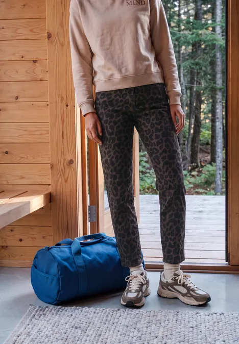 Person steht in einer Holzhütte vor offener Tür, trägt gemusterte Leggings und Sneaker, daneben blaue Reisetasche.