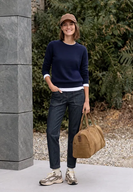 Jean taille haute à Barrel Leg, coupe ample rinsed wash Eine Frau steht draußen in marineblauem Pullover, Jeans und Sneakern, hält eine braune Reisetasche.