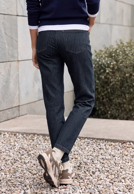 Jean taille haute à Barrel Leg, coupe ample rinsed wash Person steht im Freien, trägt dunkelblaue Nadelstreifenhose und Sneaker auf Kiesboden vor einer Mauer.