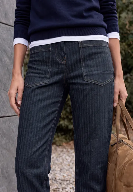 Jean taille haute à Barrel Leg, coupe ample rinsed wash Figur in dunkler Nadelstreifenjeans, Marinepullover und Tasche, steht vor einer Mauer im Freien.