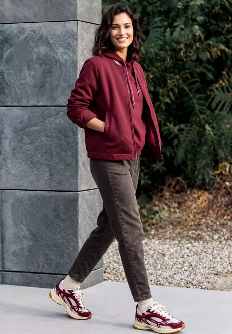 Jean « Barrel Leg » au look « garment dye » truffle brown Une femme souriante en sweat bordeaux, pantalon brun et baskets colorées appuyée contre un mur extérieur.