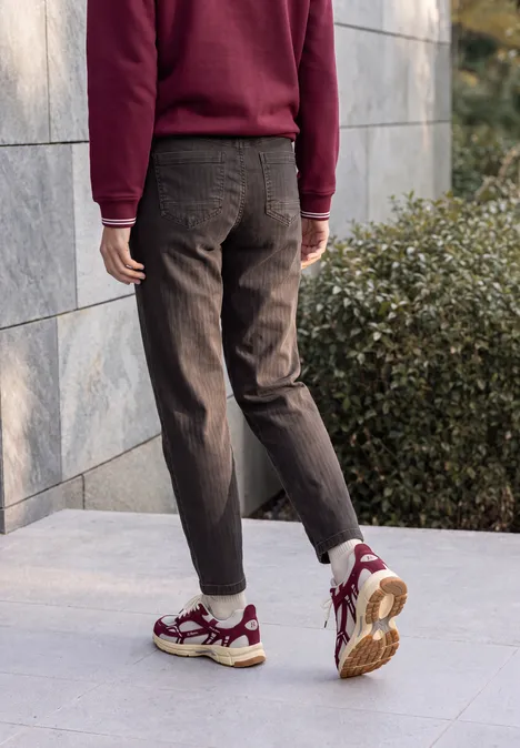 Jean « Barrel Leg » au look « garment dye » truffle brown Personne en pull bordeaux et pantalon sombre, baskets blanches à détails rouges, marche le long d'un mur en pierre.