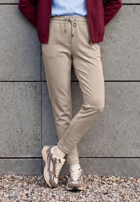 Pantalon taille mi-haute, coupe Slim Leg et style décontracté cashew beige Person steht vor Betonwand in beigen Jogginghosen, beigen Sneakern und roter Jacke