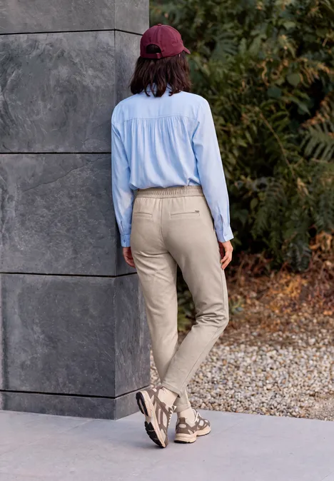 Pantalon taille mi-haute, coupe Slim Leg et style décontracté cashew beige Person mit roter Kappe, hellblauer Bluse und beigen Hosen läuft an Mauer und herbstlichem Laub vorbei.