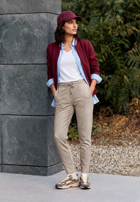 Pantalon taille mi-haute, coupe Slim Leg et style décontracté cashew beige Frau lehnt an Hauswand im Freien, trägt burgunderrote Jacke, hellblaue Bluse, weißes Top, beige Hose und Sneaker.