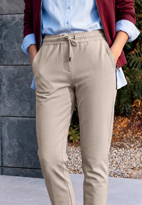 Pantalon taille mi-haute, coupe Slim Leg et style décontracté cashew beige Teilansicht einer Person in beigen Jogginghosen mit Kordelzug, blauem Hemd und rotem Sakko.