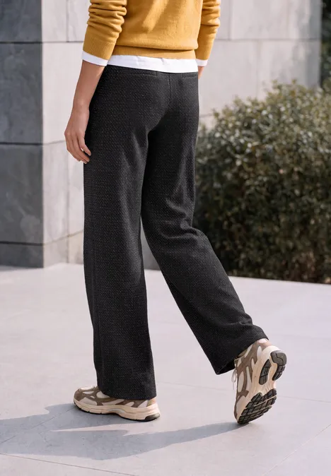Persoon in losse donkere broek en sneakers loopt langs een moderne gevel.