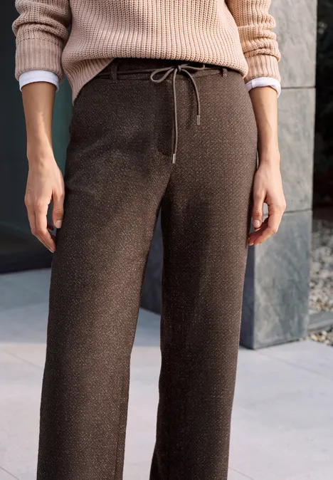 Person in weitem braunen Strickhose mit Tunnelzug und beigem Pullover vor Steinwand.