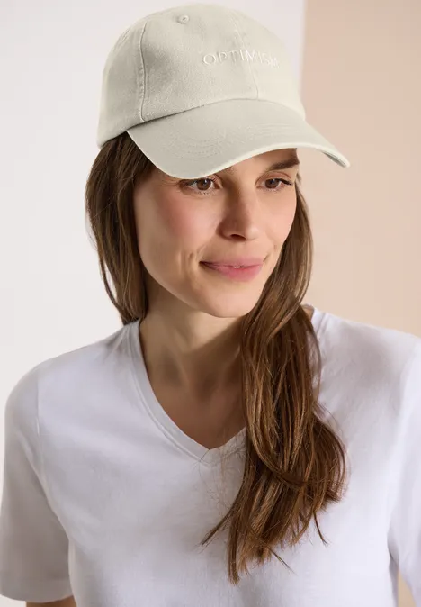 Person mit weißem T-Shirt und weißer Baseballcap vor zweifarbigem, hellem Hintergrund.