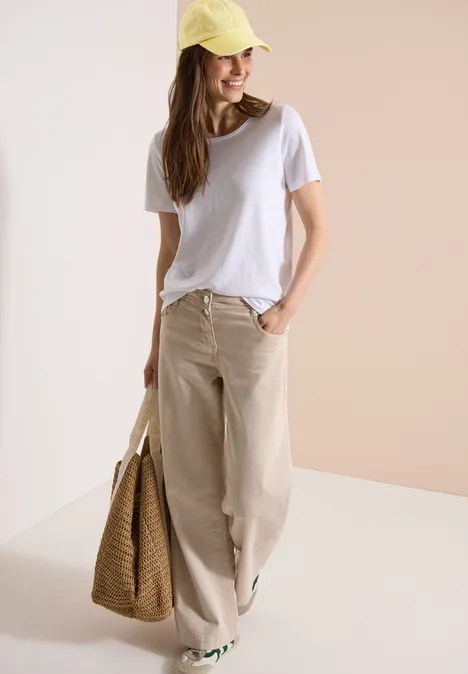 Frau in weißem T-Shirt, weitem beigen Stoffhosen und gelber Mütze mit Strohtasche.
