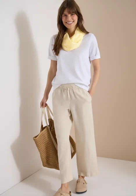 Frau steht vor Wand, trägt weißes T-Shirt, beige weite Hose, gelbes Halstuch, Korbtasche und Sandalen.