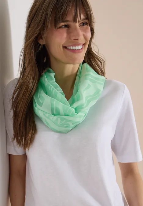 Een vrouw draagt een lichtgroene nekwarmer en wit T-shirt, leunend tegen een muur.