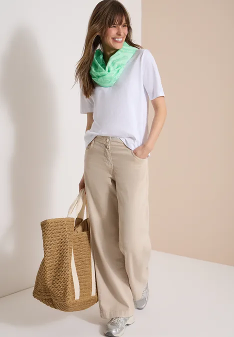 Vrouw met wit T-shirt, brede beige broek, groene sjaal, houdt een strooien tas vast.