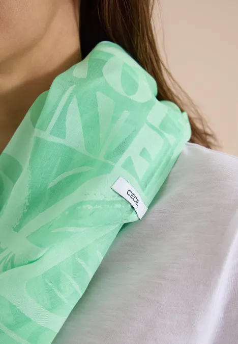 Lichtgroene sjaal met wit motief en GCDS-label, nonchalant over één schouder gelegd, gecombineerd met een wit shirt.