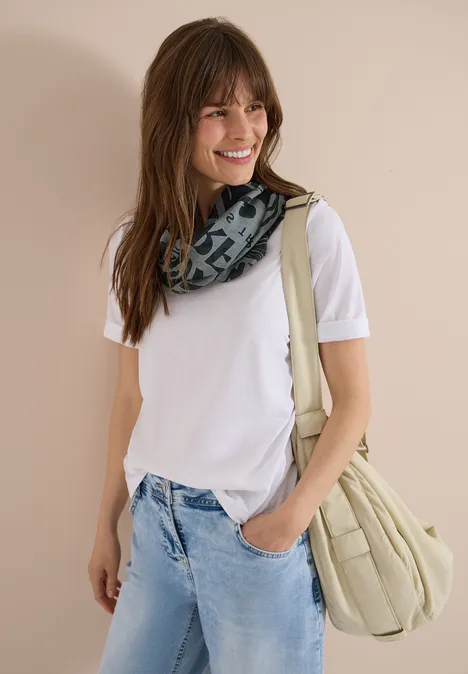 Jeune femme en t-shirt blanc, jean et écharpe à motifs, sac beige sur l’épaule devant un mur clair.
