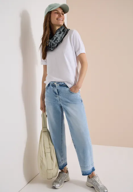 Femme en t-shirt blanc, jean bleu, casquette, foulard, grand sac matelassé et baskets.