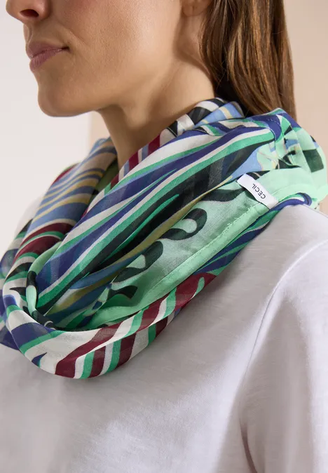 Une femme porte un foulard infini aux rayures vertes, bleues et blanches sur un t-shirt blanc.