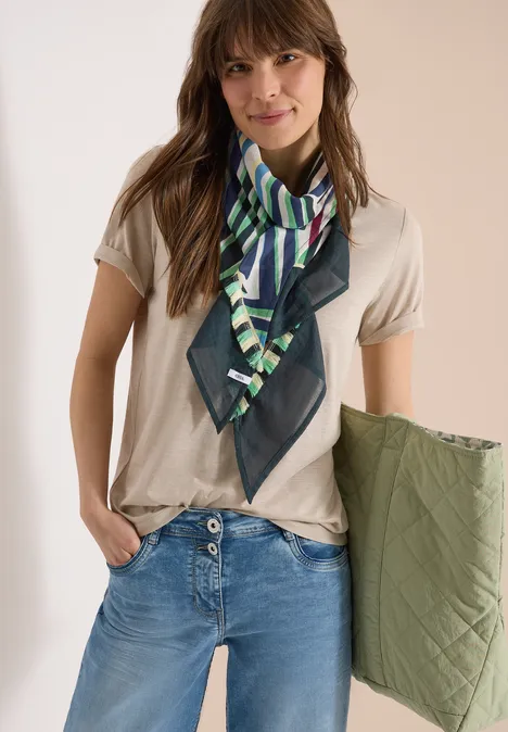 Femme en t-shirt beige, jean bleu, écharpe verte à motifs, tenant une veste matelassée kaki