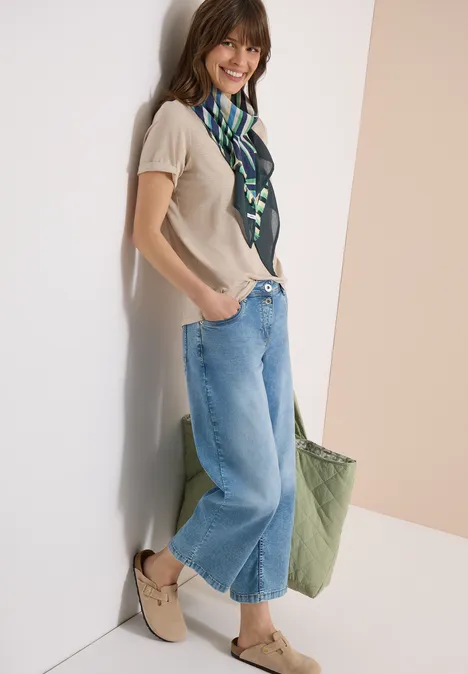 Une femme appuyée contre un mur porte un top beige, un jean ample, un foulard vert et un sac vert.