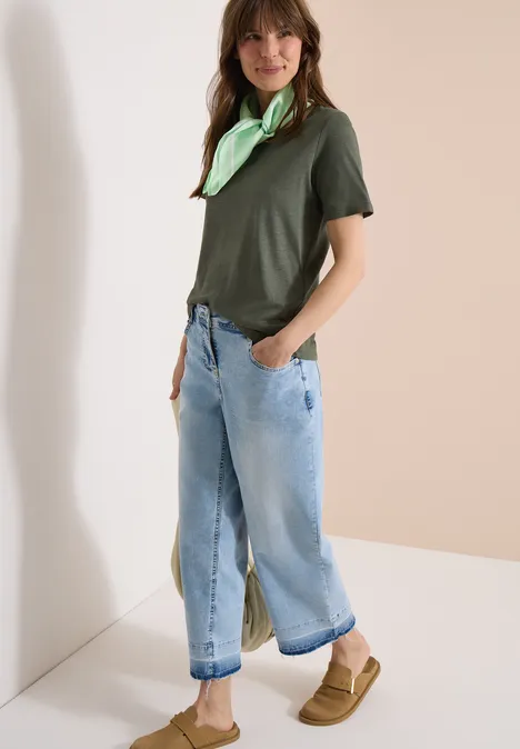 Vrouw in groen T-shirt, lichtgroene sjaal, wijde opgerolde jeans en beige loafers.