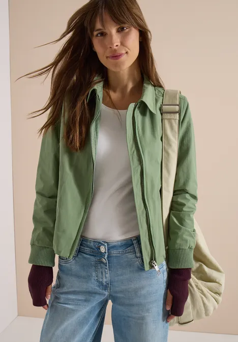 Eine Frau trägt grüne Jacke, weißes Top, Jeans, auberginefarbene Handschuhe und eine beige Umhängetasche.