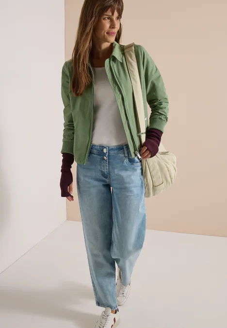 Eine lächelnde Frau in grüner Jacke, hellem Top, Jeans und weißen Sneakern trägt eine Umhängetasche.