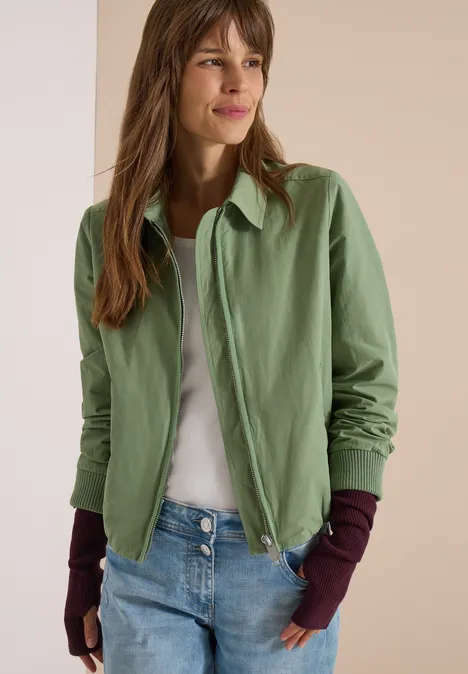 Frau trägt hellgrüne Jacke, weißes Shirt, blaue Jeans und lila Handstulpen.