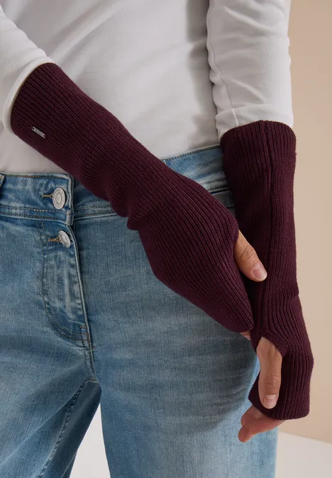 Weinrote, fingerlose Strickhandschuhe, getragen über hellblauer Jeans und weißem Oberteil.