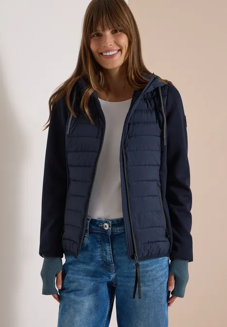 Frau in lässiger, dunkelblauer Steppjacke mit Kapuze, weißem Shirt und Jeans, lächelt entspannt.