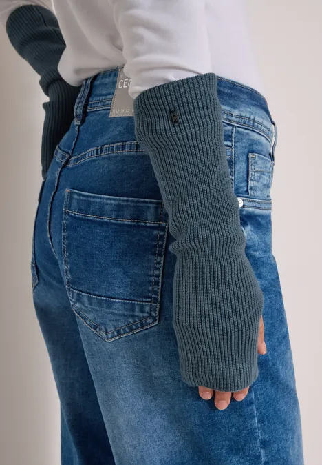 Eine Person trägt graue Strick-Handstulpen und Jeans, dazu ein weißer Pullover.