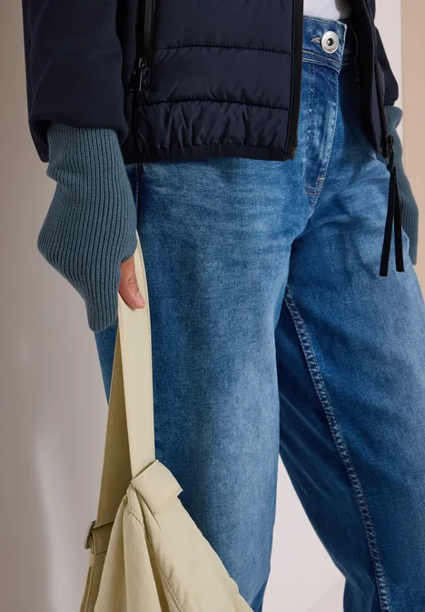 Nahaufnahme einer Person in Jeans, schwarzer Steppjacke und fingerlosen Handschuhen mit beiger Tasche.