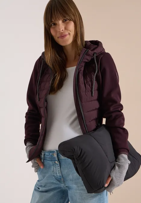 Frau mit langen braunen Haaren trägt eine burgunderfarbene Jacke, weiße Bluse, Jeans und graue Handschuhe.